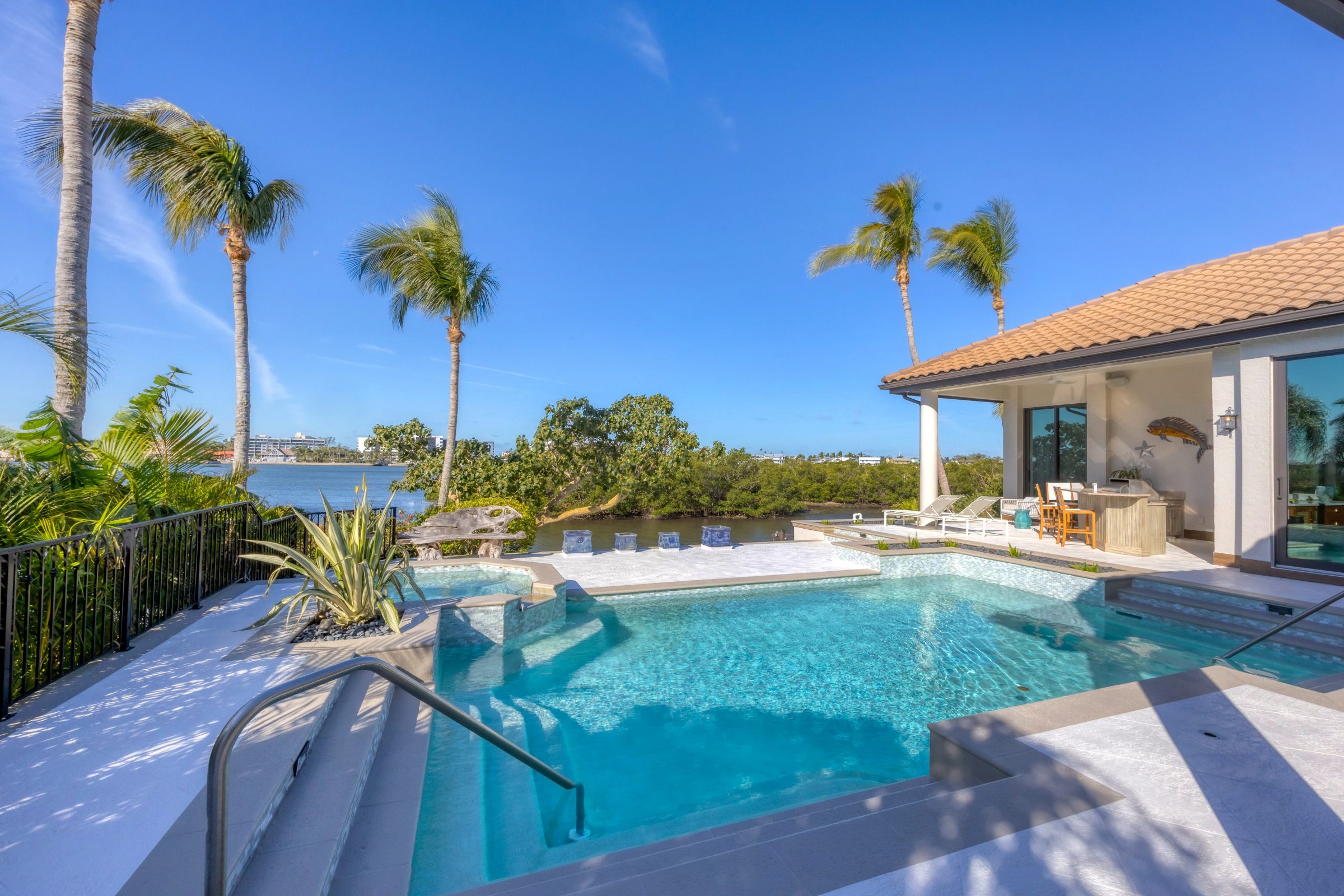 An elegant pool with a spa, overlooking the ocean with palm trees and a modern patio seating area.