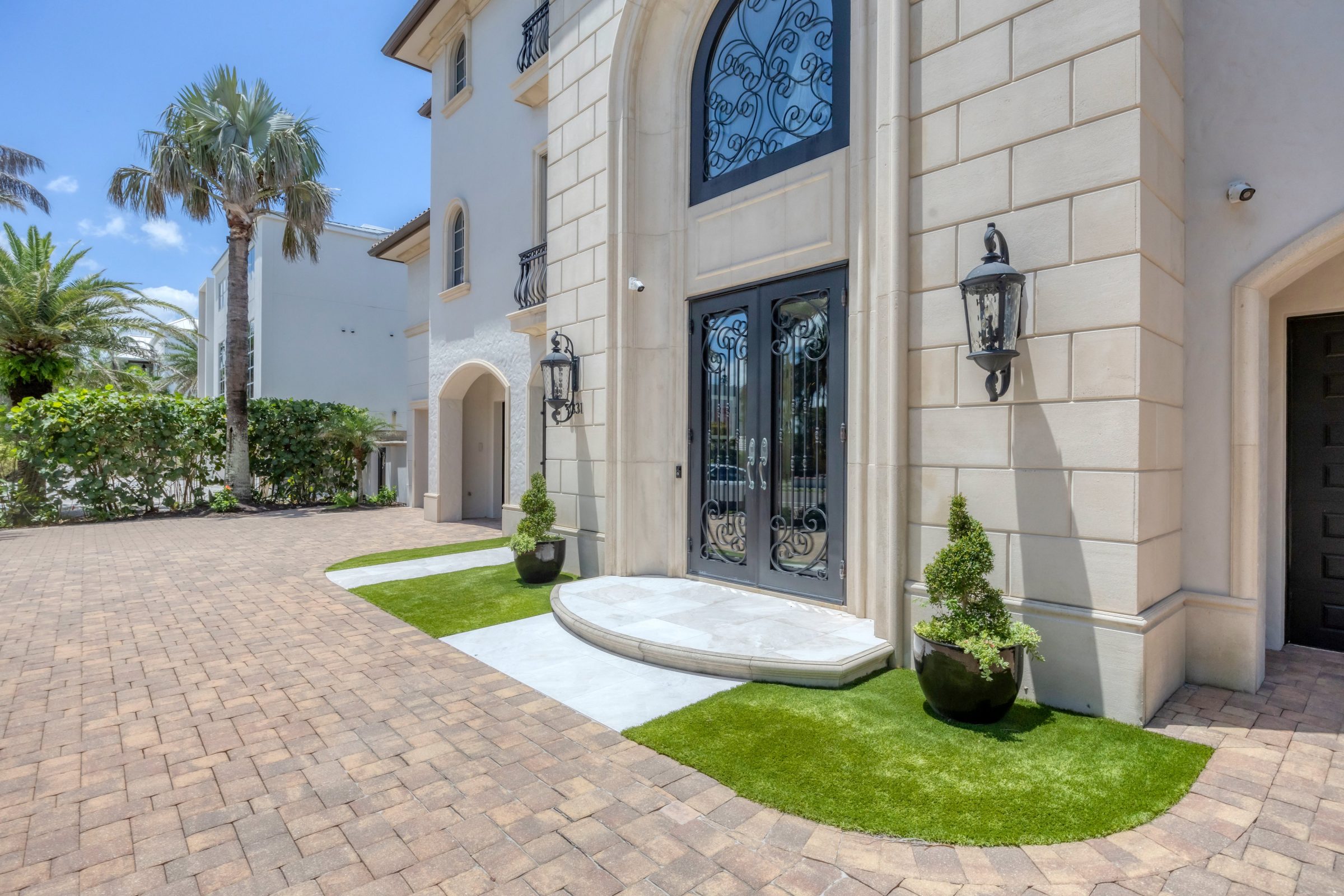 Ornate wrought iron double doors, stone walls, potted greenery, and a paved driveway.