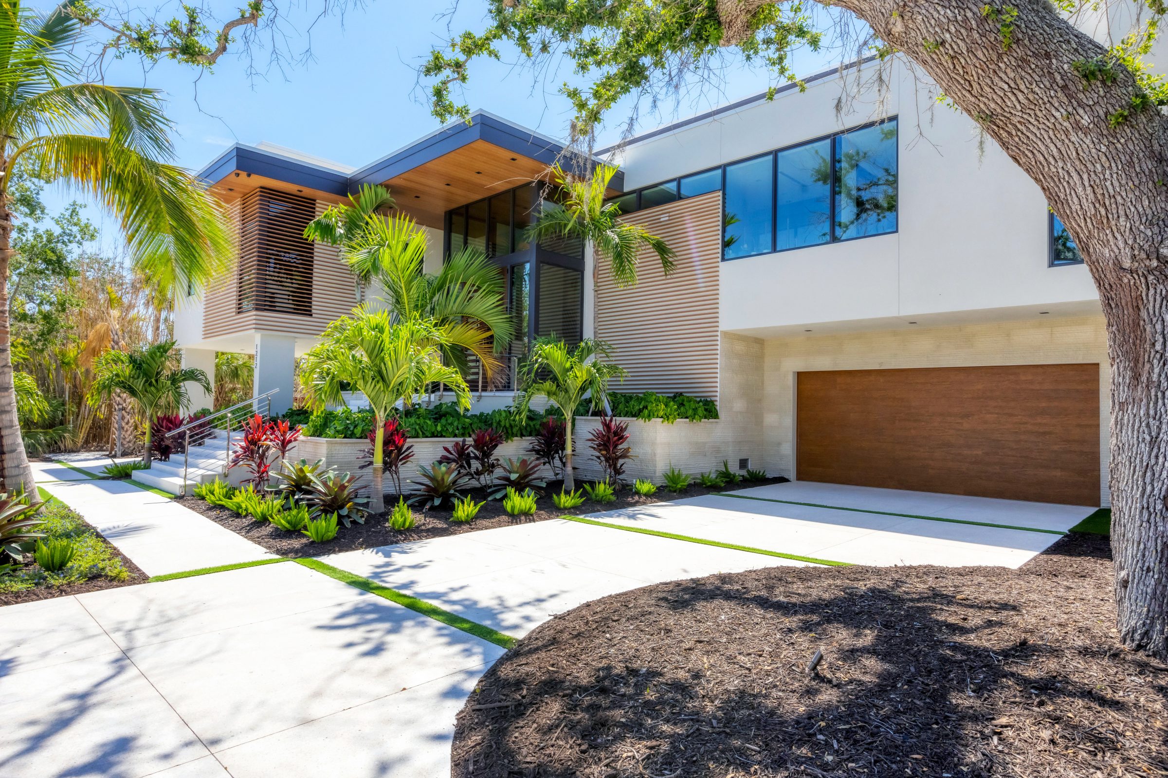 Sleek modern facade with wood accents, tall windows, and lush plantings of palms, bromeliads, and red foliage.