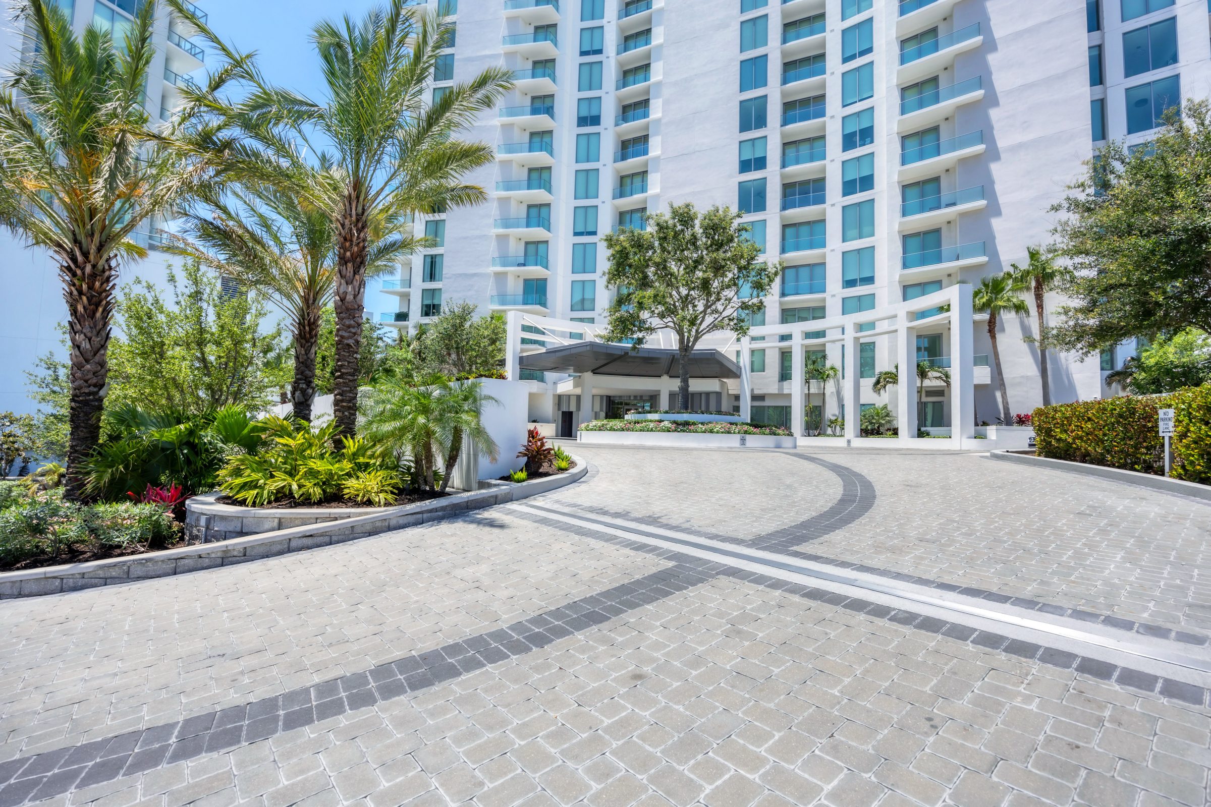 Circular driveway with pavers, tall palms, manicured greenery, and a modern high-rise building behind.